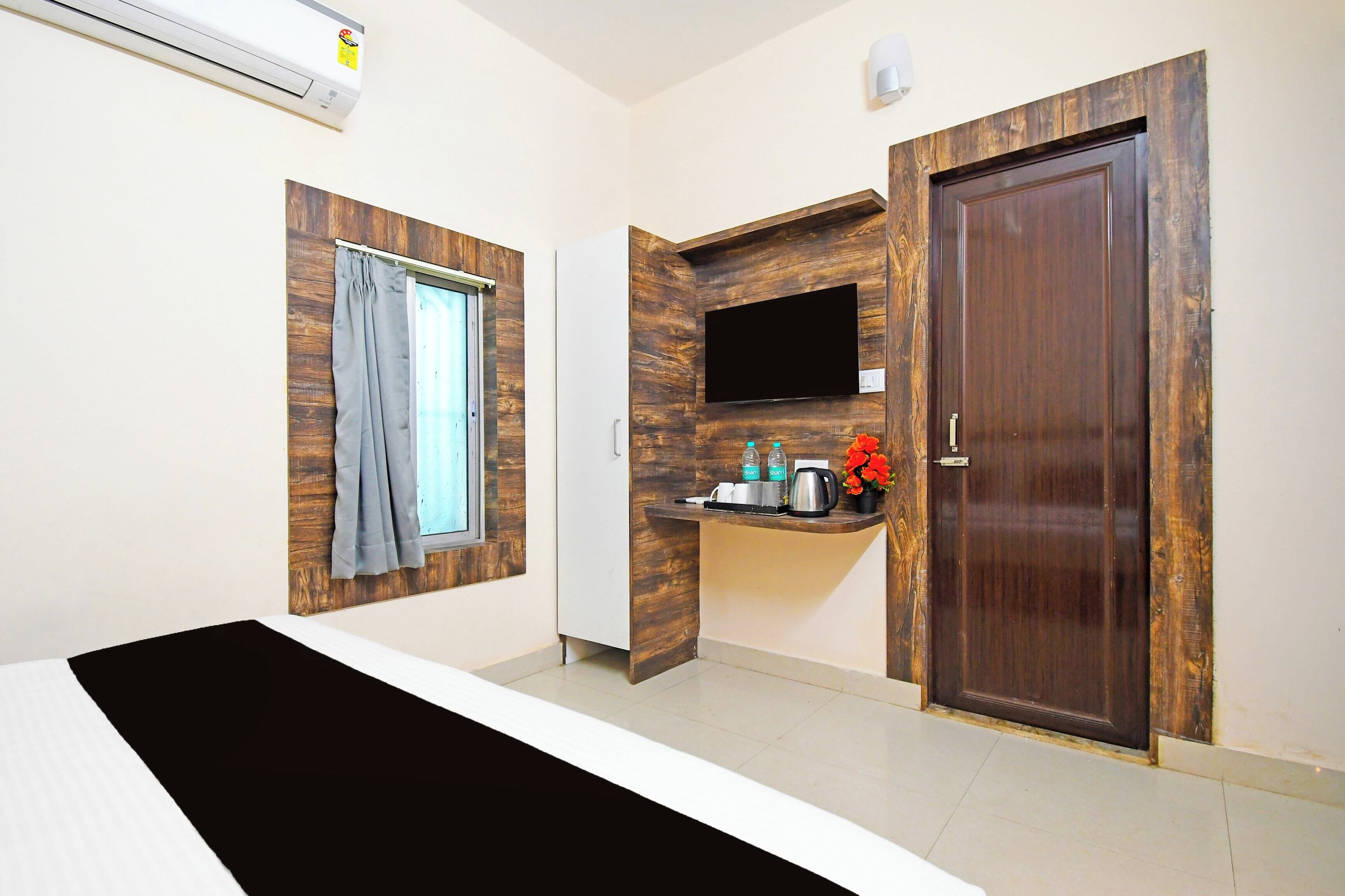 Une chambre d’hôtel avec un lit, une fenêtre avec des rideaux, un téléviseur fixé au mur, une petite étagère avec des articles de toilette et une porte en bois.