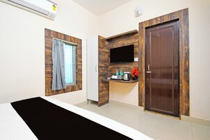 Une chambre d’hôtel avec un lit, une fenêtre avec des rideaux, un téléviseur fixé au mur, une petite étagère avec des articles de toilette et une porte en bois.