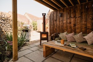 A cozy outdoor seating area with a stone wall, a wooden table, and a built-in wood stove. - Boutique Cotswolds Cottage near Soho Farmhouse (Duns Tew)