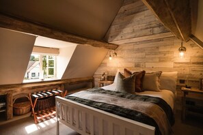 A cozy attic bedroom with a sloped ceiling, wooden walls, a bed with pillows, a nightstand, a lamp, and a window with a view. - Boutique Cotswolds Cottage near Soho Farmhouse (Duns Tew)