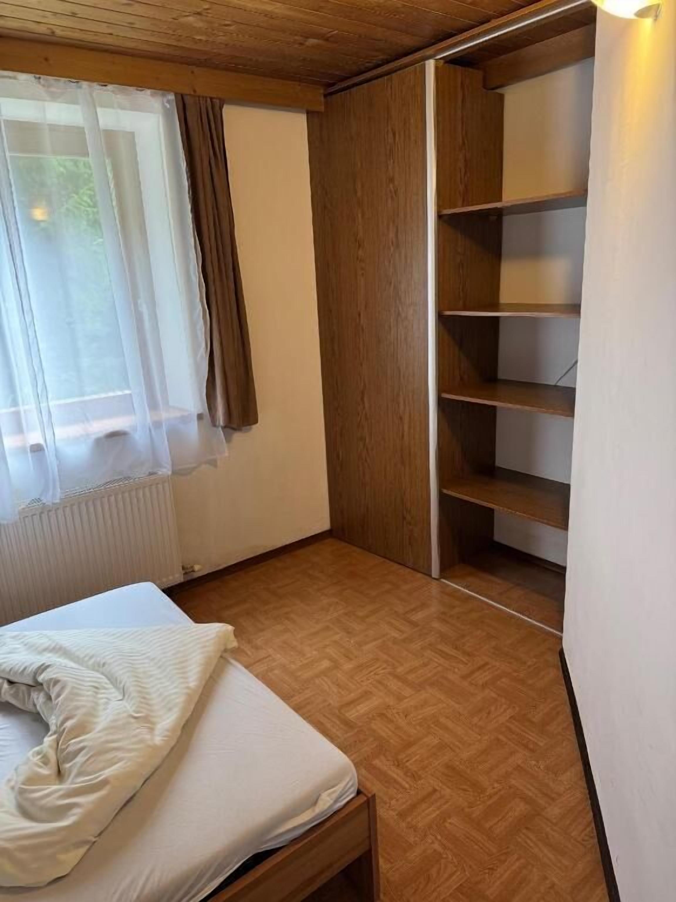 Una habitación con una cama, una ventana con cortinas translúcidas, un radiador y un armario de madera con estantes.