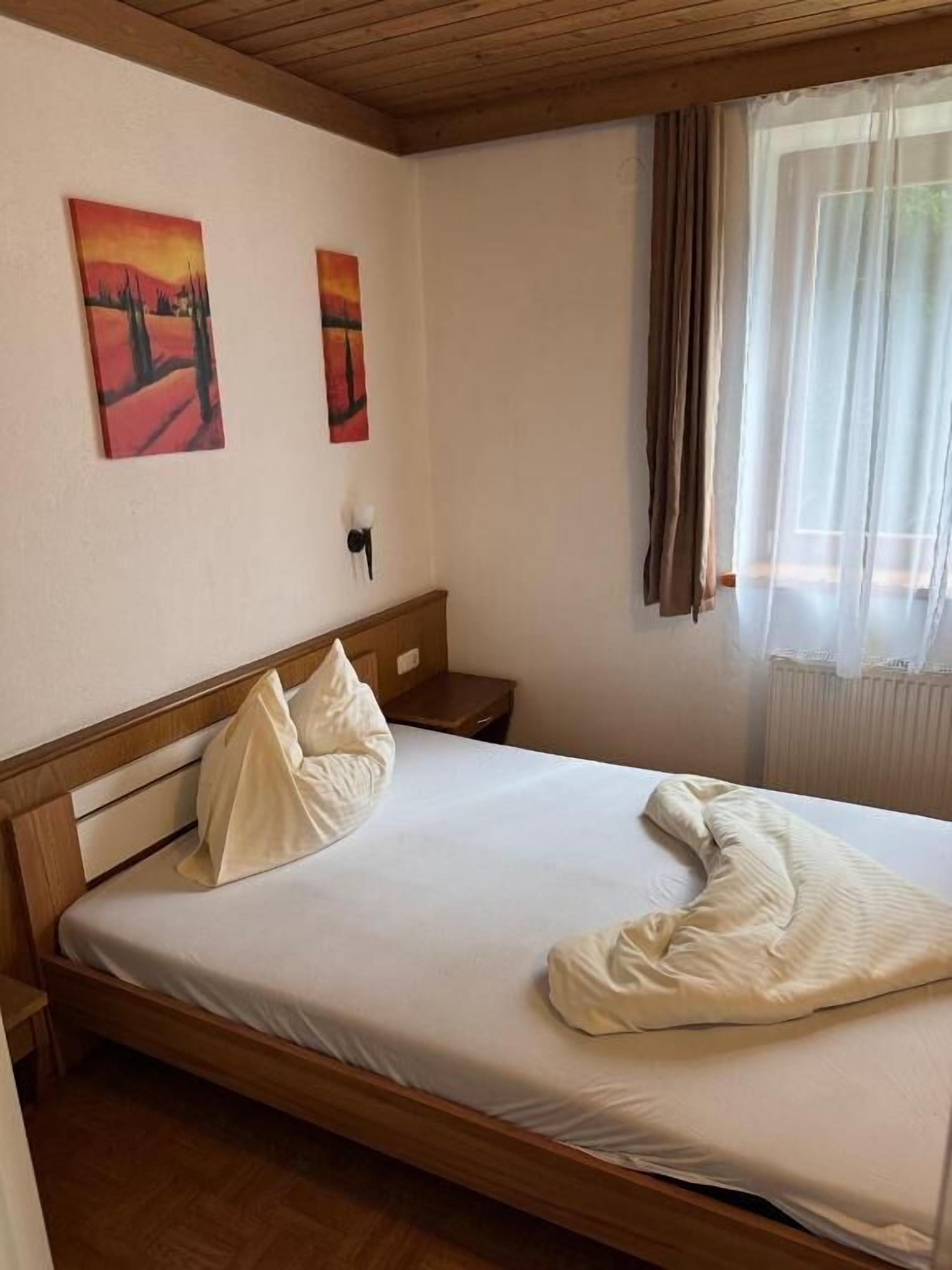 A single bed with a wooden headboard, two wall-mounted paintings, a bedside lamp, and a window with curtains.