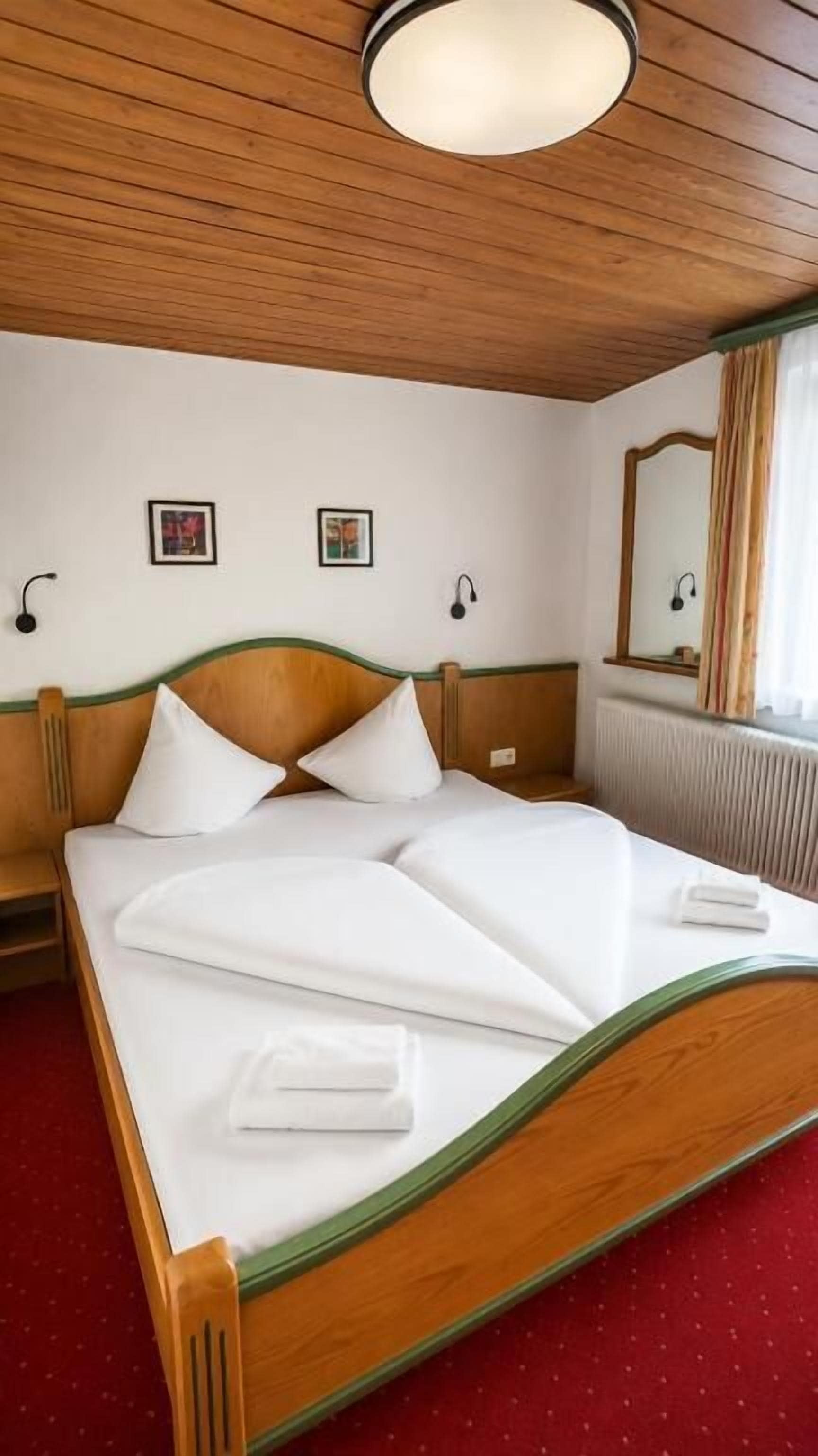 Una habitación de hotel con una cama de madera, ropa de cama blanca, una alfombra roja y un espejo grande.