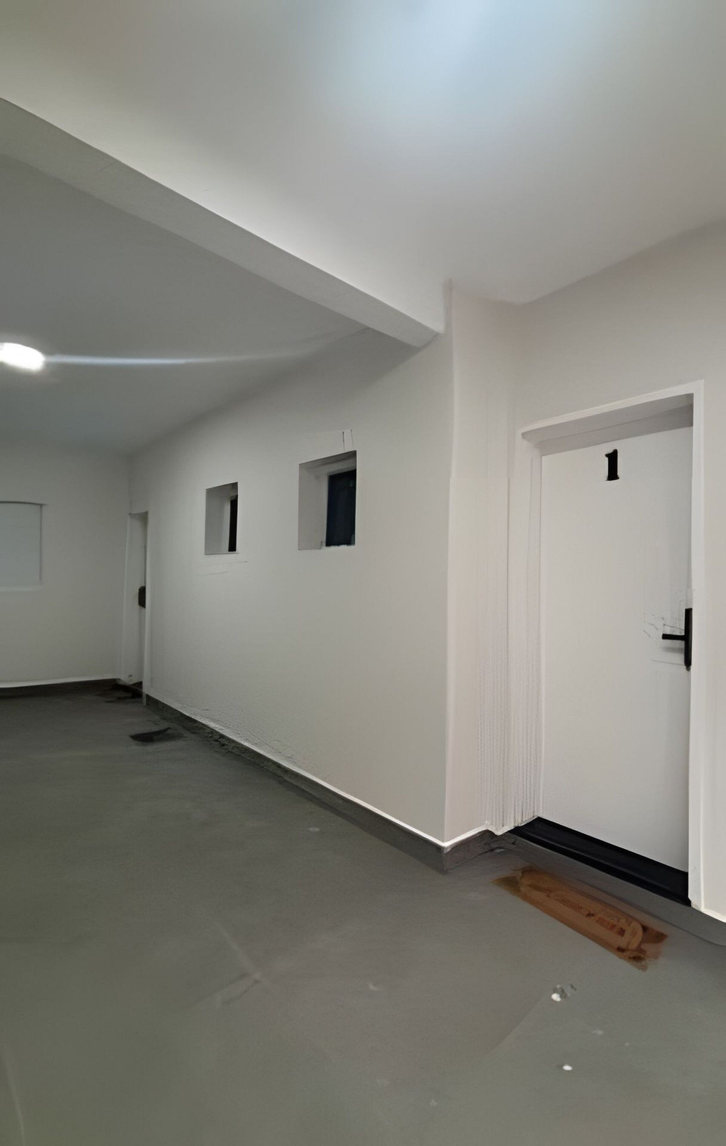 Um quarto espaçoso e vazio, com paredes brancas e chão cinzento, destacando uma porta com placa de número e uma pequena janela.