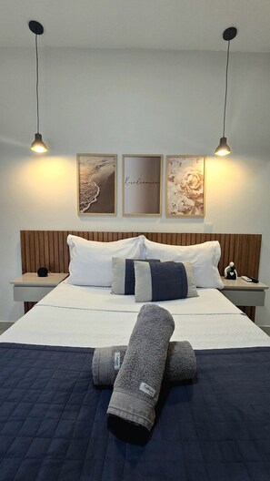 Un lit bien fait, recouvert d’une couette bleue, avec deux oreillers et une couverture enroulée. De chaque côté du lit, il y a une table de chevet sur laquelle sont posés une lampe et une petite figurine. Au-dessus du lit, trois œuvres d’art encadrées ornent le mur.