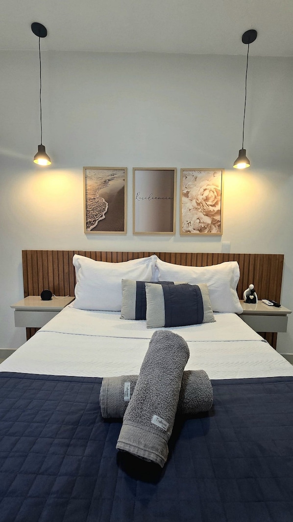 A neatly made bed with a blue quilt, two pillows, and a rolled-up blanket. There are two bedside tables with a lamp and a small figurine on each. Above the bed, there are three framed artworks on the wall.
