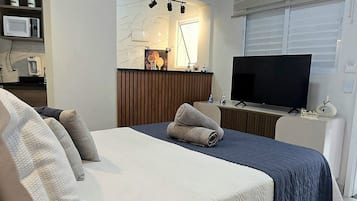 Quarto de hotel com uma cama, televisão, ar condicionado e uma pequena cozinha.