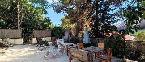 An outdoor patio area with wooden furniture, including tables and chairs, surrounded by trees and a wooden fence.