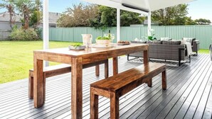 An outdoor patio with a wooden table and benches, a covered seating area with cushions, and a view of a green fence and trees in the background. - Periwinkle - perfect base for a beachside getaway with family and pets (Culburra Beach)