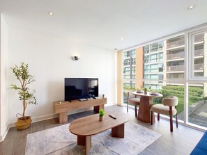 A modern living room with a flat-screen TV, wooden furniture, and a large window offering a view of a building. - Fully Serviced One Bed Apartment near London Excel (Greater London)