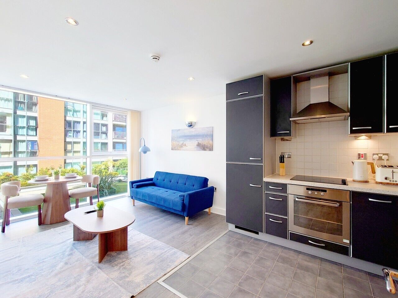 A modern kitchen with dark cabinets, a built-in oven, and a dining area with a blue sofa and round table.