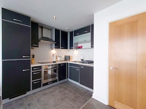 A modern kitchen with dark cabinetry, a built-in oven, and a microwave. - Fully Serviced One Bed Apartment near London Excel (Greater London)