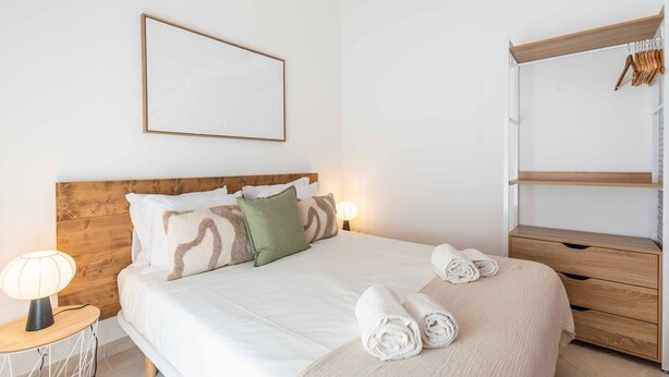 Ein ordentlich eingerichtetes Schlafzimmer mit einem Holz-Kopfteil, einem Bett mit weißer Bettwäsche, einer Nachttischlampe und einem Kleiderschrank.
