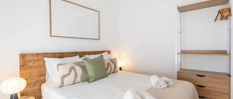 Quarto bem organizado com cabeceira de madeira, cama com roupa de cama branca, abajur e guarda-roupa suspenso.