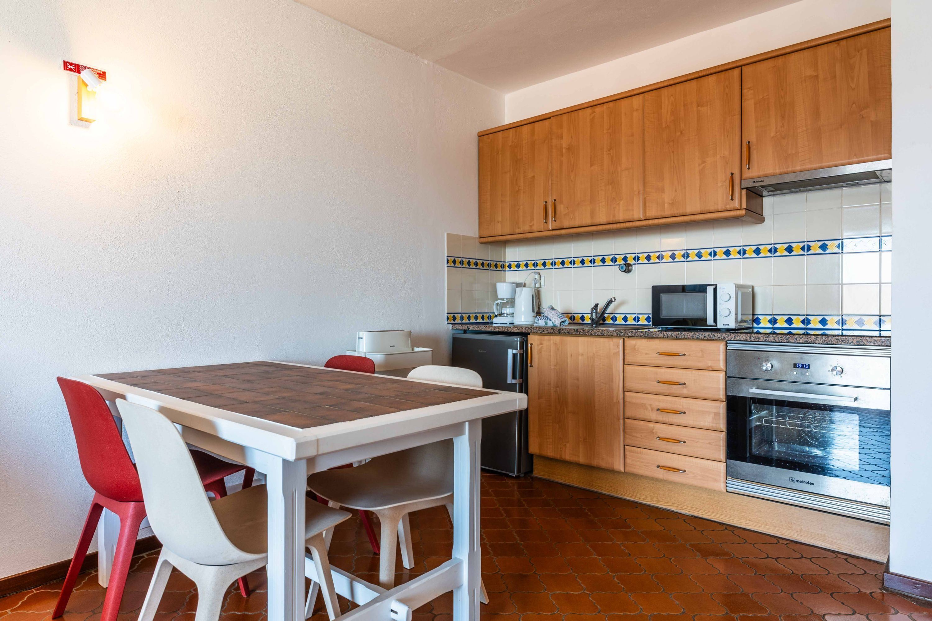 Una cocina compacta con armarios de madera, salpicadero de azulejos, horno integrado y un comedor con mesa y sillas.