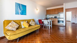 A living room with a sofa, a dining table with chairs, a kitchen area with cabinets and appliances, and a tiled floor. - Apartamento T1 Vistamar Centro Hist Rico Albufeira (Albufeira)