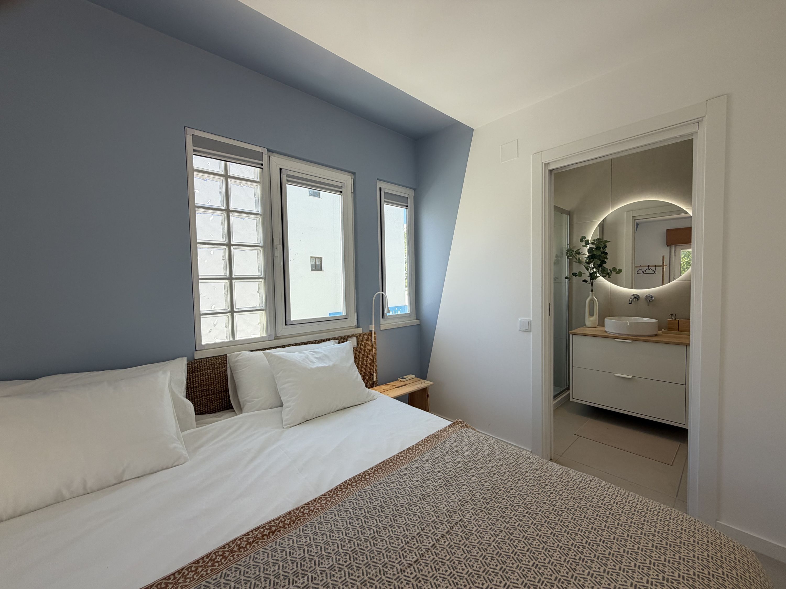 A bedroom with a bed, a nightstand, a mirror, and a bathroom with a sink and mirror.