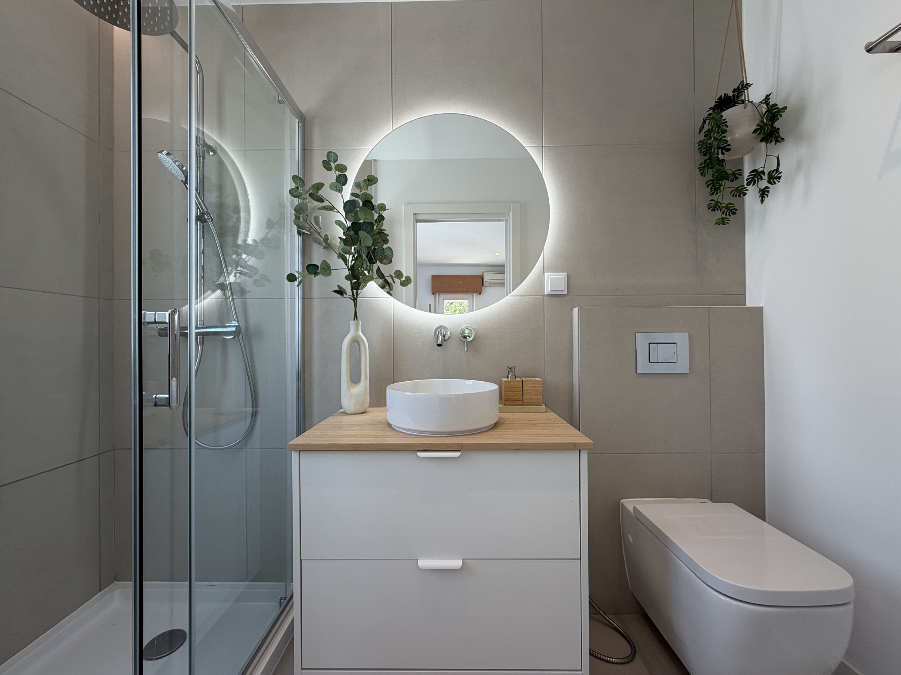 Un bagno moderno con box doccia in vetro, uno specchio rotondo, un mobile bagno in legno con lavandino e un water.