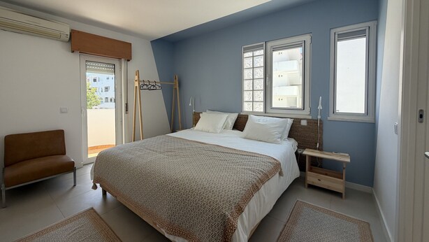 A bedroom with a bed, a nightstand, a chair, and a window with a view of buildings. - Central Town 2-bedroom Classy Apartment Albufeira (Albufeira)