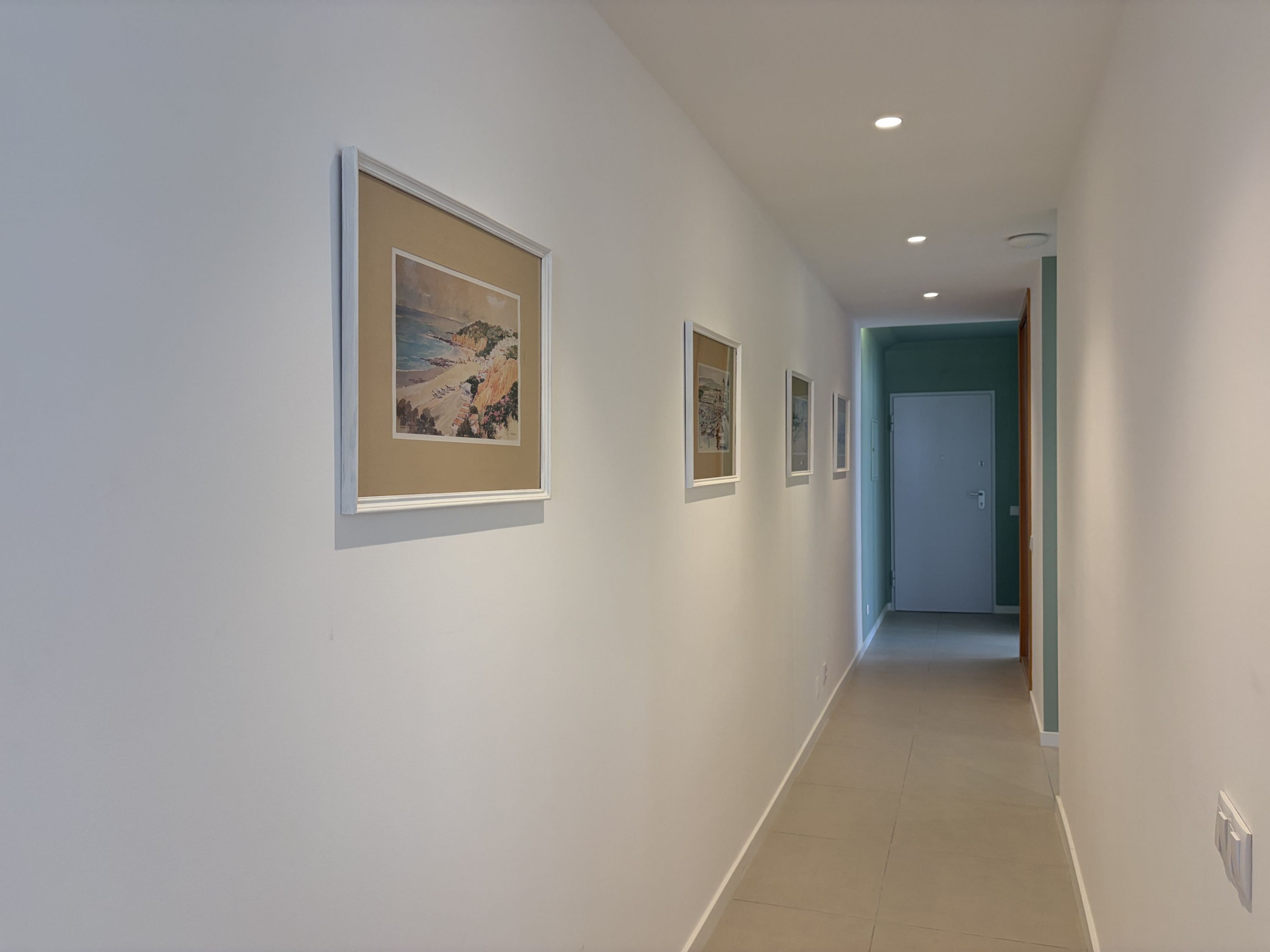 A hallway with framed pictures on the walls, a door at the end, and recessed lighting.