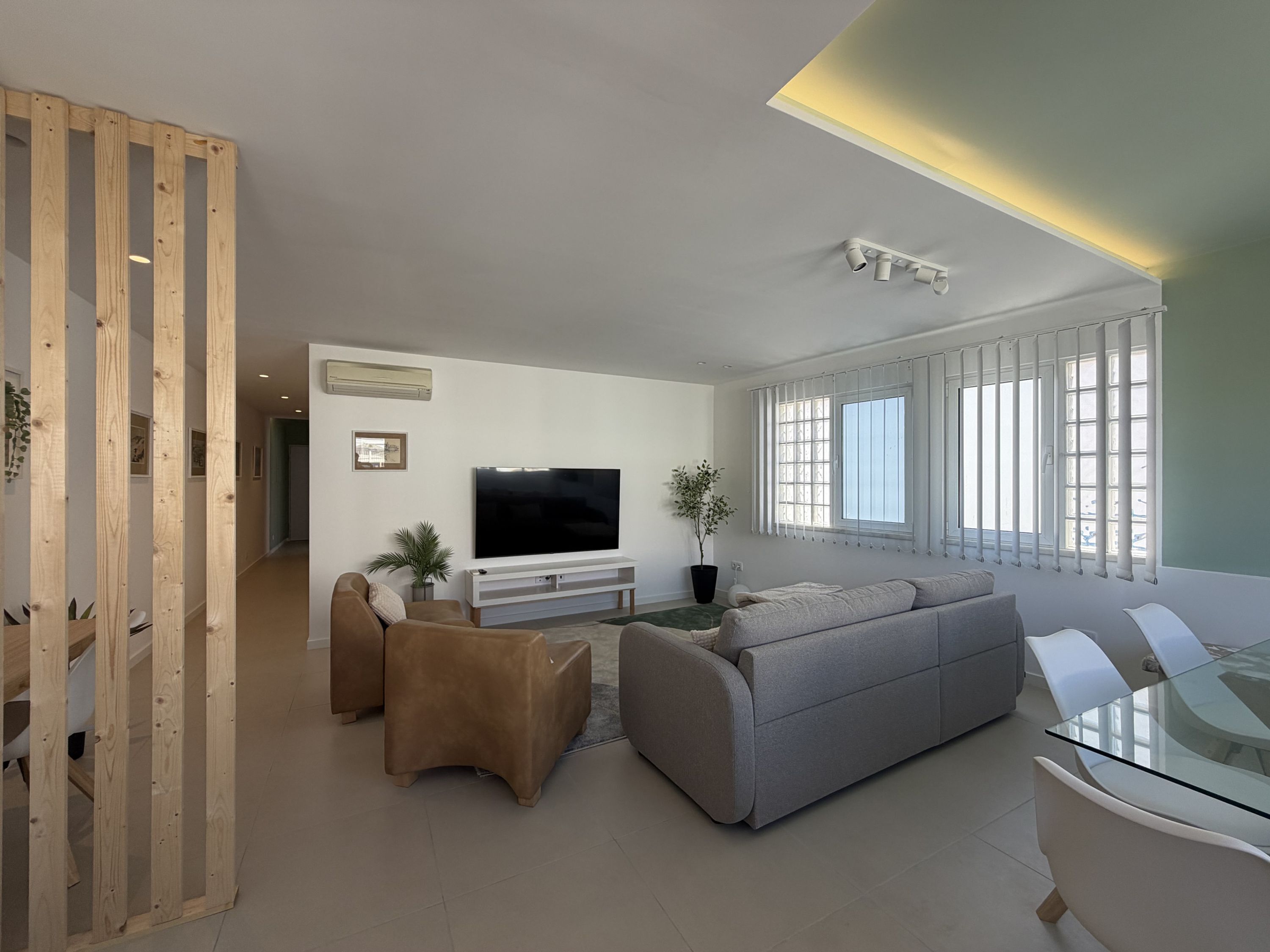 A modern living room with a flat-screen TV, a grey sofa, a glass coffee table, and a wooden partition.