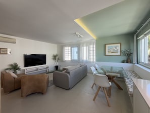 A modern living room with a flat-screen TV, a sofa, a dining table with chairs, and a large window with blinds. - Central Town 2-bedroom Classy Apartment Albufeira (Albufeira)