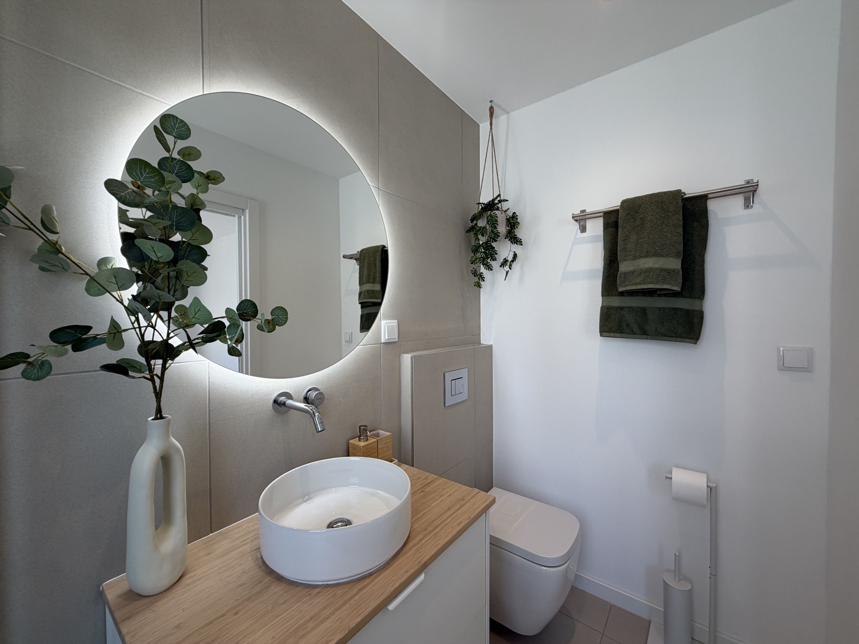 A modern bathroom with a round mirror, a white sink, a toilet, and a towel rack.