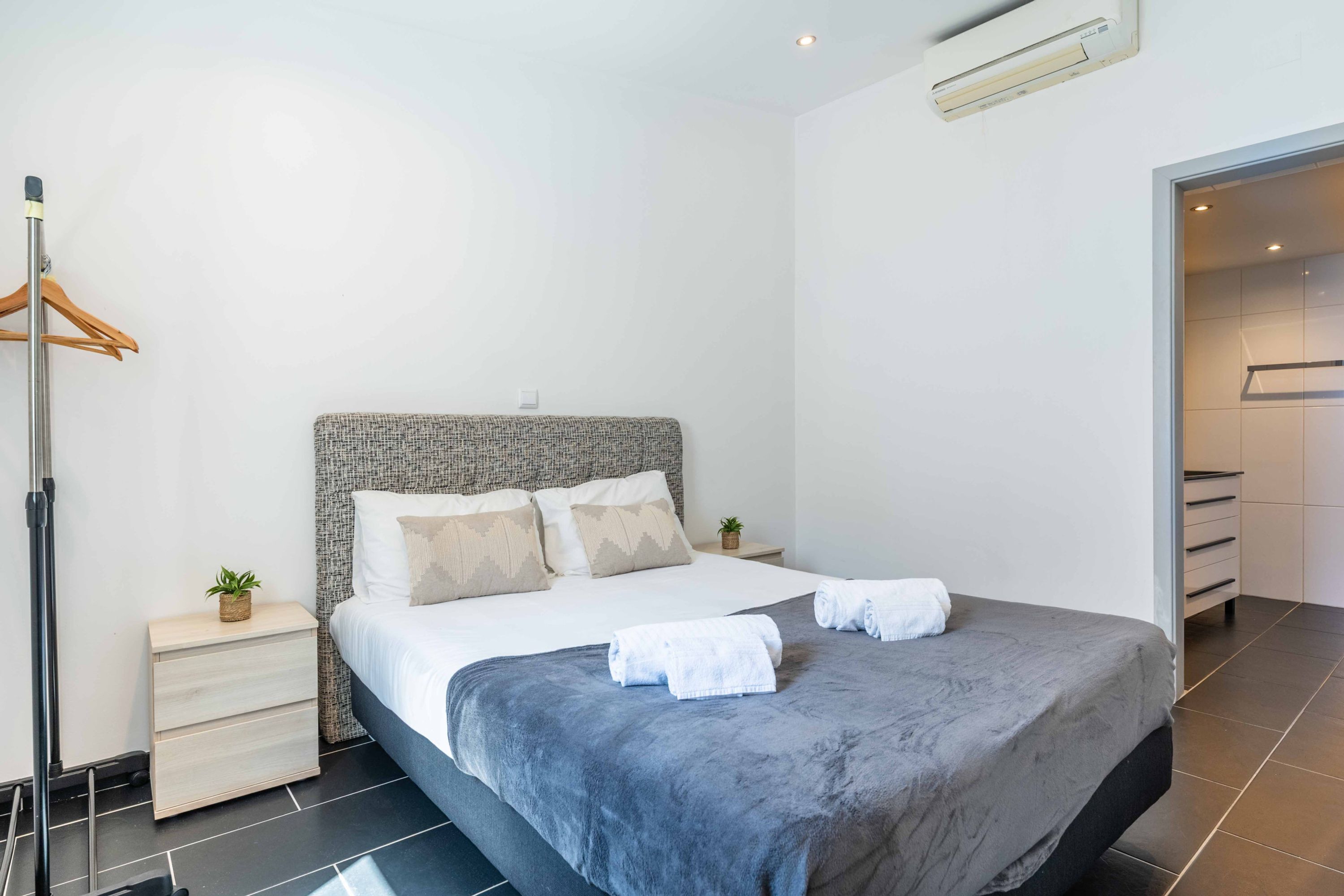 Quarto de hotel moderno com uma cama grande, uma mesa de cabeceira com abajur, uma pequena planta e um guarda-roupa.