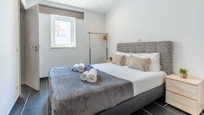 A modern bedroom with a large bed, a bedside table, a nightstand, a window, and a door. - Geneve Old Town T3 Apartment (Albufeira)