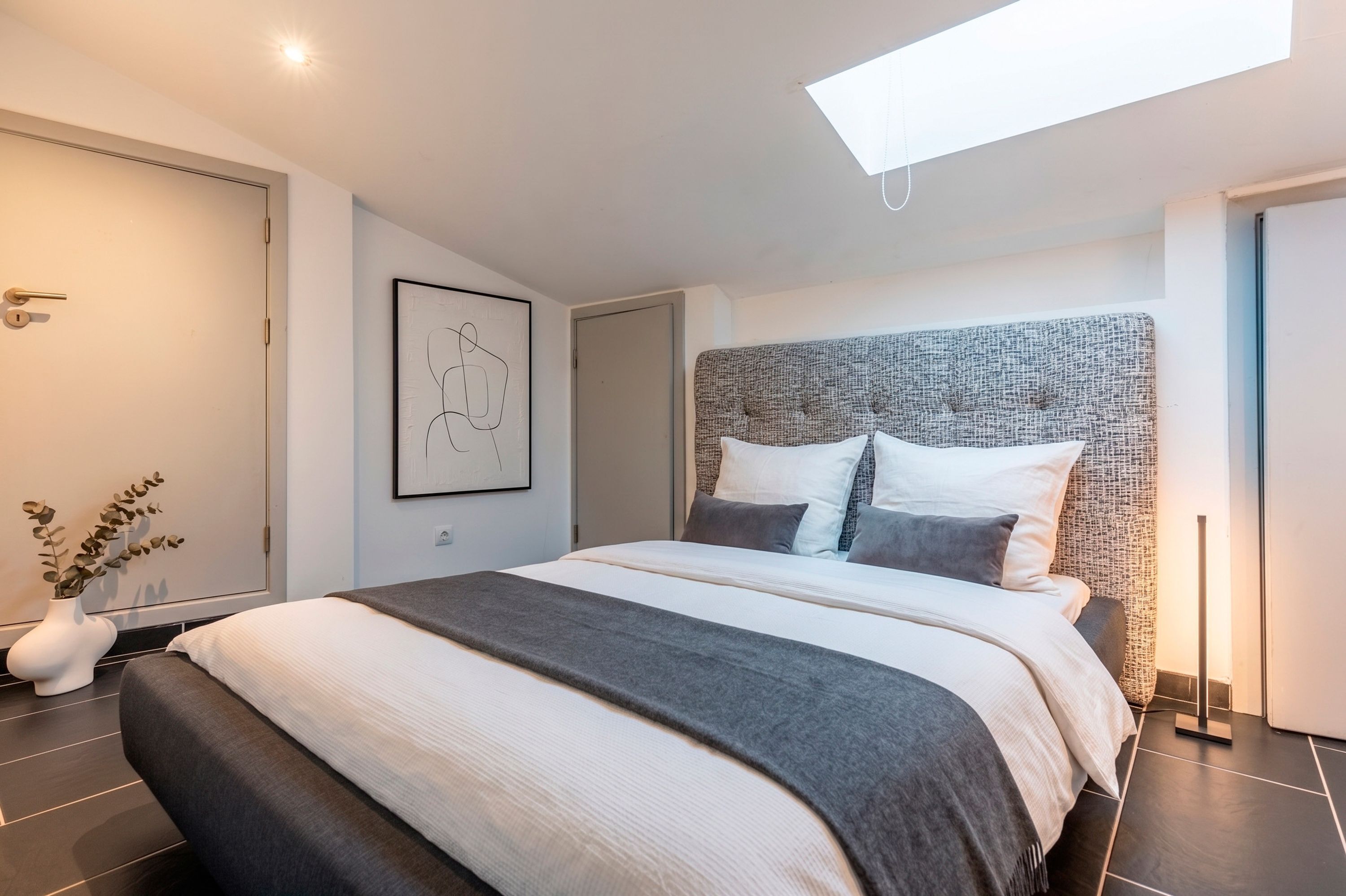A modern bedroom with a large bed, a headboard with a textured pattern, a framed abstract artwork on the wall, and a skylight above the bed.