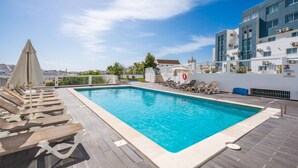Uma piscina com espreguiçadeiras e guarda-sóis, cercada por edifícios e um céu claro.