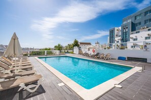 Ein Schwimmbecken mit Liegestühlen und Sonnenschirmen, umgeben von Gebäuden und einem klaren Himmel.