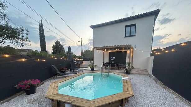 A rectangular swimming pool with a wooden deck, surrounded by gravel, and a patio area with outdoor furniture.