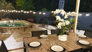 An outdoor dining area with a wooden table set for two, surrounded by chairs, and a hot tub in the background.