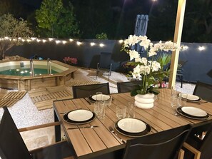 An outdoor dining area with a wooden table set for two, surrounded by chairs, and a hot tub in the background.