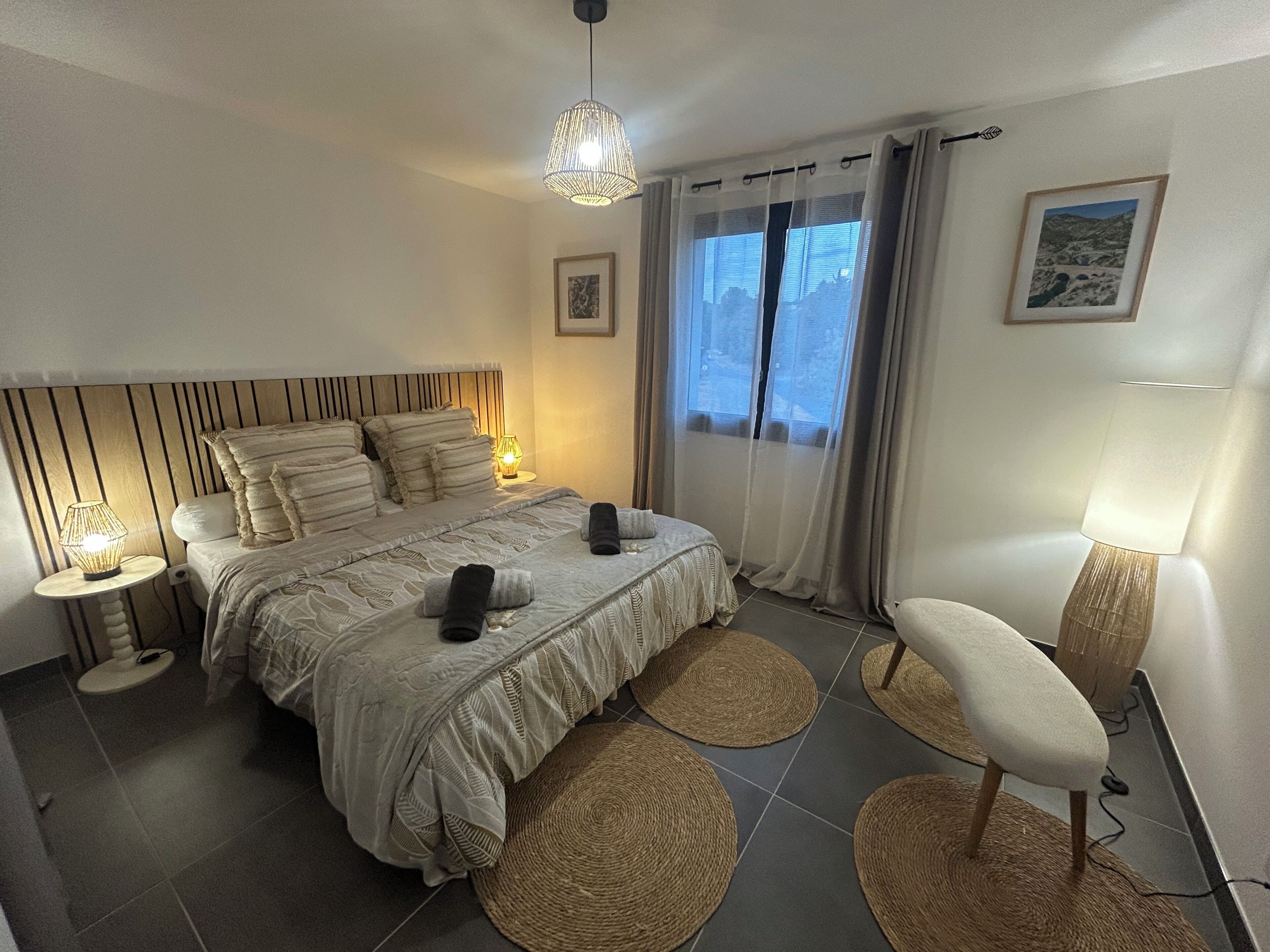 Ein modernes Schlafzimmer mit einem großen Bett, Nachttischlampen, Blick ins Freie durch ein Fenster und einem Sessel mit Lampe.