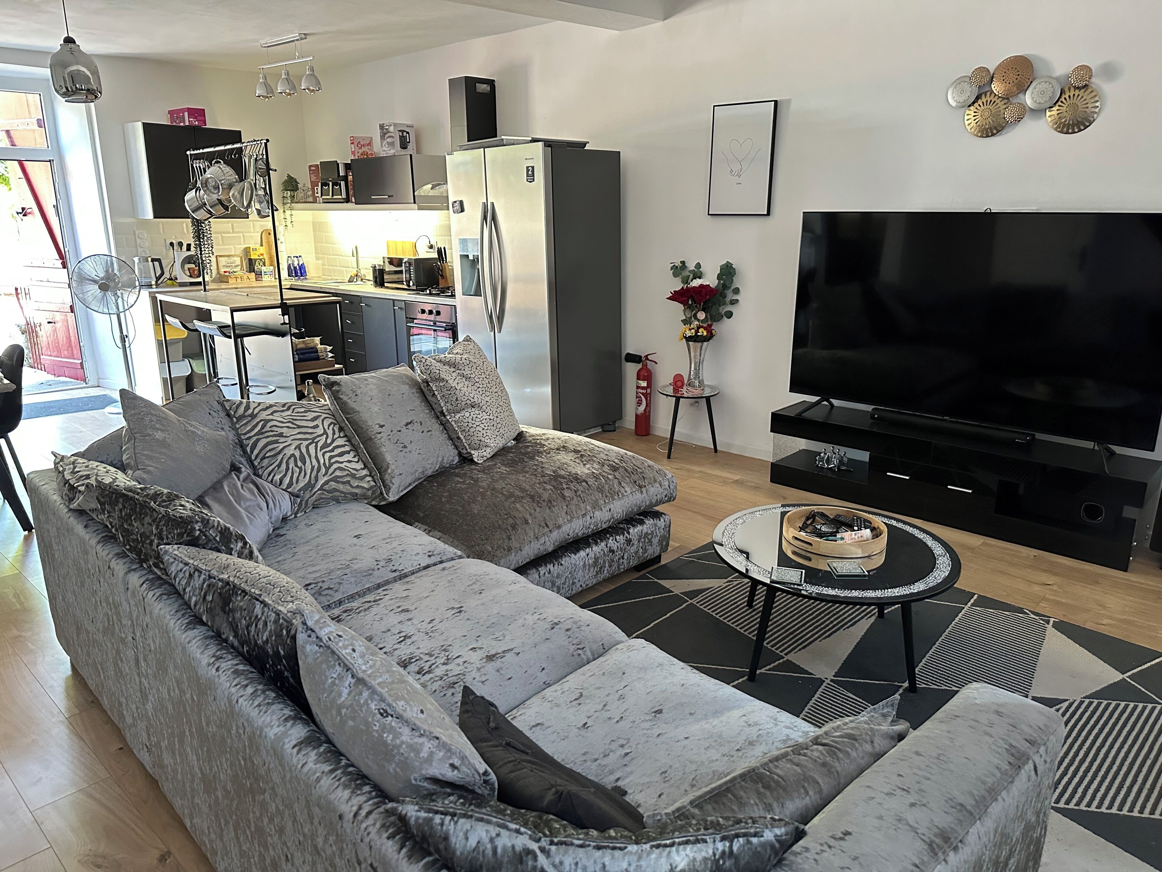 Una sala moderna con un sofá seccional gris, un televisor de pantalla plana y una cocina al fondo.