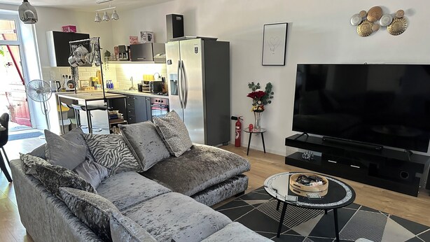 Una sala moderna con un sofá seccional gris, un televisor de pantalla plana y una cocina al fondo.