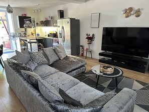A modern living room with a grey sectional sofa, a flat-screen TV, and a kitchen area in the background. - Located in the heart of the village, at the foot of the Château de Bonneval. (Coussac-Bonneval)