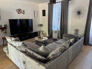 Ein modernes Wohnzimmer mit einem großen Flachbildfernseher, einem grauen Sofa mit gemusterten Kissen, einem kleinen runden Couchtisch und einer Wanddekoration.