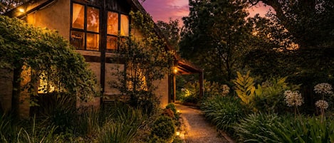 Una casa rústica con fachada de madera, un ventanal visible y un sendero de jardín que conduce a una estructura de madera.