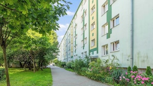 Un sendero conduce a un edificio de varios pisos con fachada verde y blanca, rodeado de abundante follaje y plantas con flores.