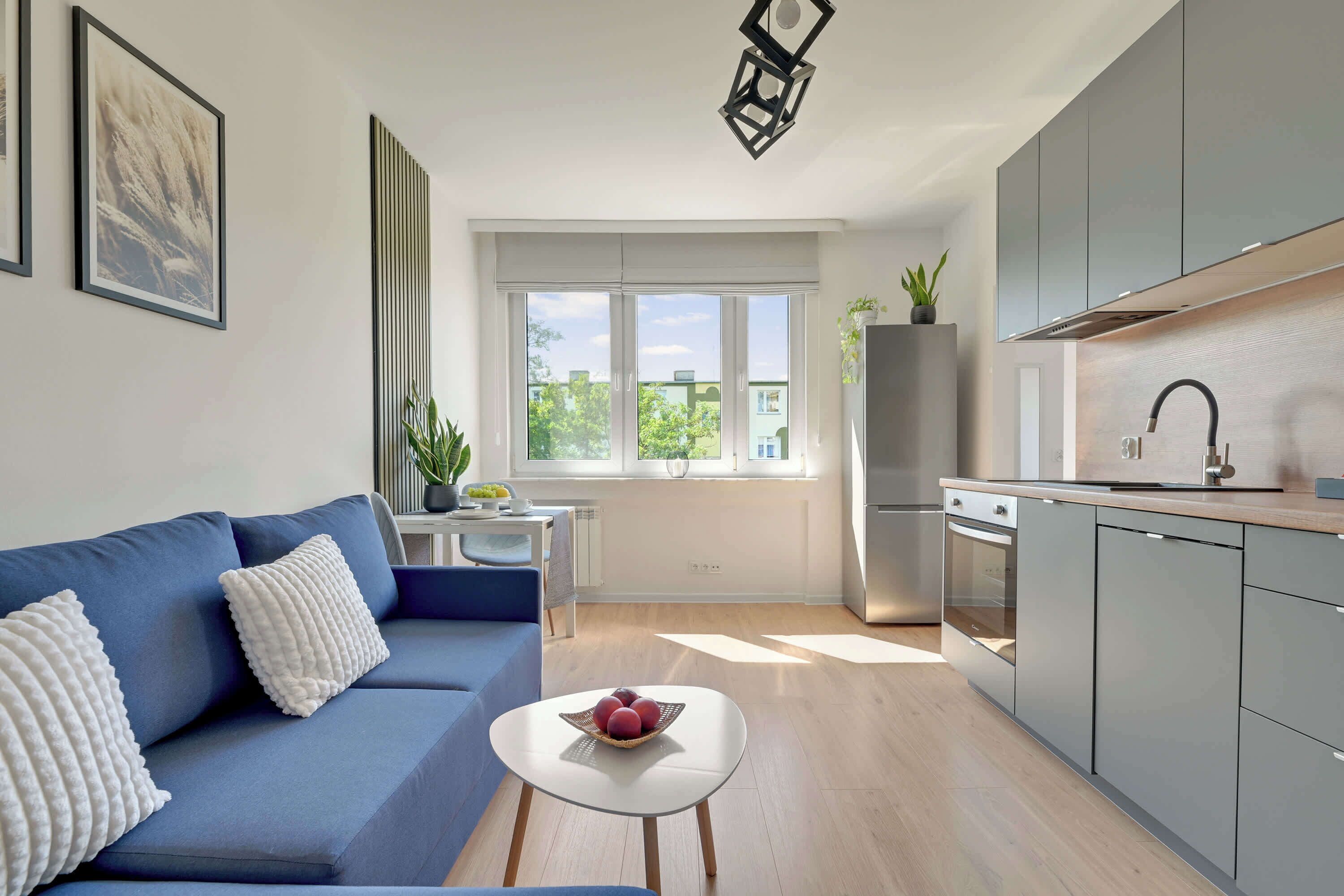 A modern kitchen with a built-in oven, a stainless steel refrigerator, and a grey cabinet. There is a blue sofa with white cushions and a small white table with a fruit bowl.