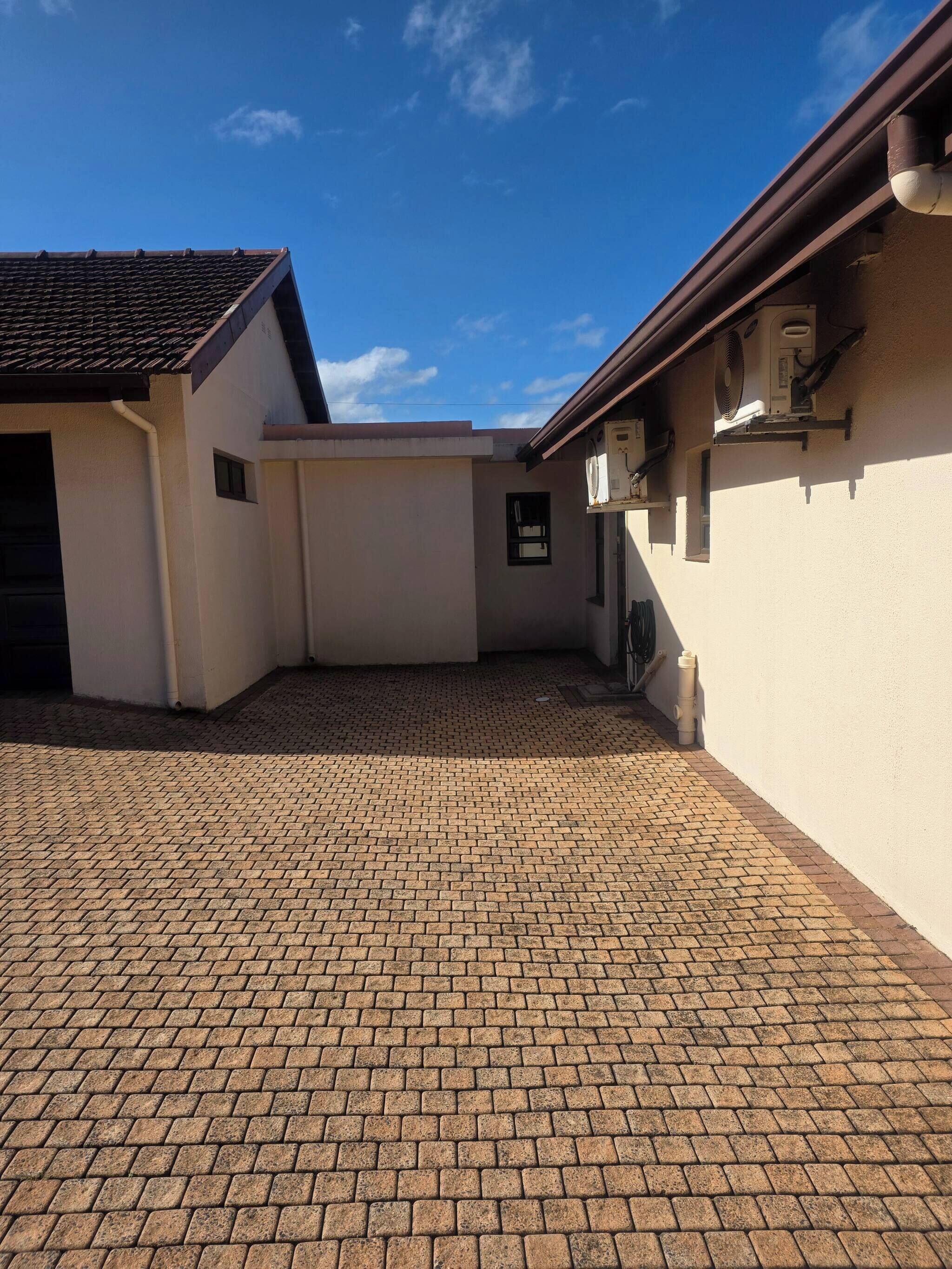 Um pátio pavimentado com telhado de azulejos, um edifício branco com uma pequena janela e um aparelho de ar condicionado fixado na parede.