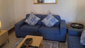 Ein Wohnzimmer mit einem blauen Sofa, gemusterten Kissen, einem Holztisch, einer Fernbedienung und einer kleinen, schwarzen Ablagefläche.