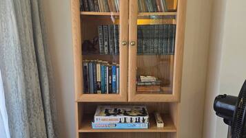 A wooden bookshelf with books and board games, a patterned tablecloth, and a black fan.