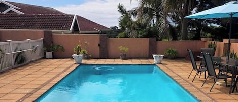 A rectangular swimming pool with a tiled edge, surrounded by a patio area with chairs and a table, and a white fence.