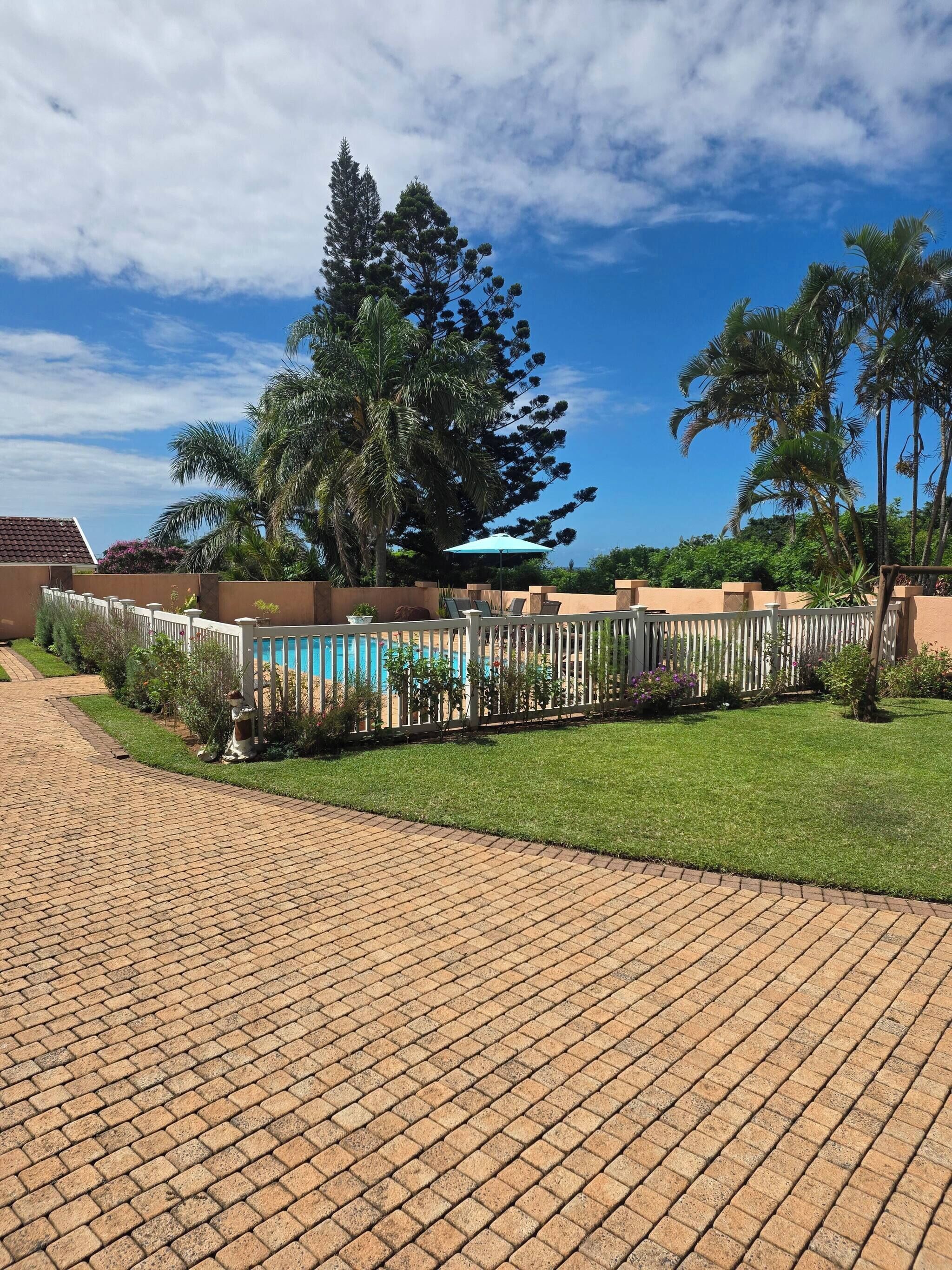 Un camino pavimentado que conduce a una zona de piscina con valla, rodeada de follaje y palmeras.