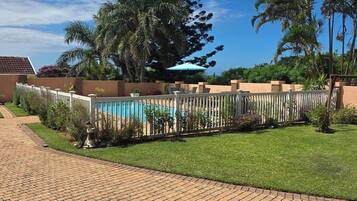 Un camino pavimentado que conduce a una zona de piscina con valla, rodeada de follaje y palmeras.
