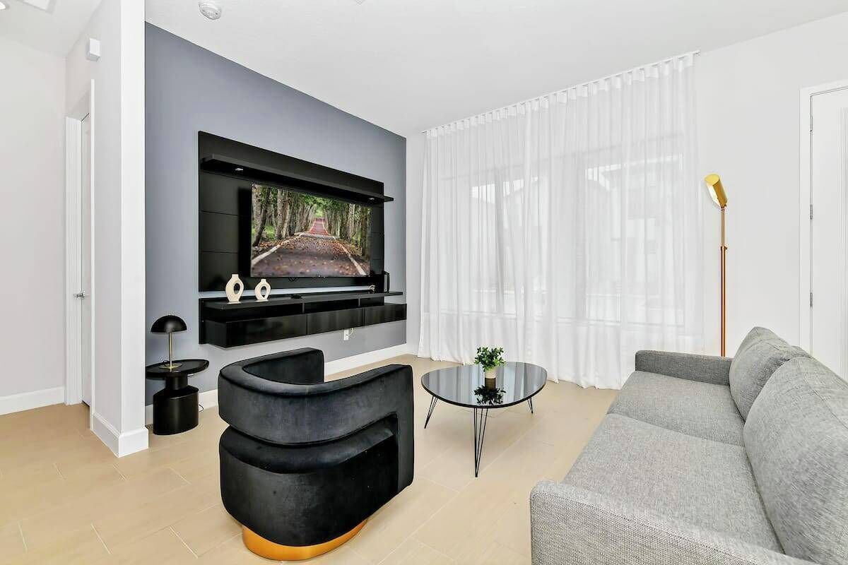 Una sala de estar moderna con un televisor de pantalla plana montado en la pared, un sofá gris, un sillón negro decorativo, una pequeña mesa redonda de centro y una lámpara de pie.